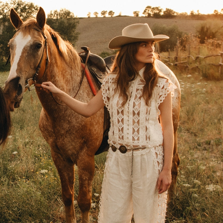 Woman wearing The Rodeo Smooth Shadow — a beige wool fedora with a classic western silhouette, styled with a white lace two-piece beside a chestnut horse at sunset. For Old Habits Die Hard, an Amsterdam-based hat-wear brand.
