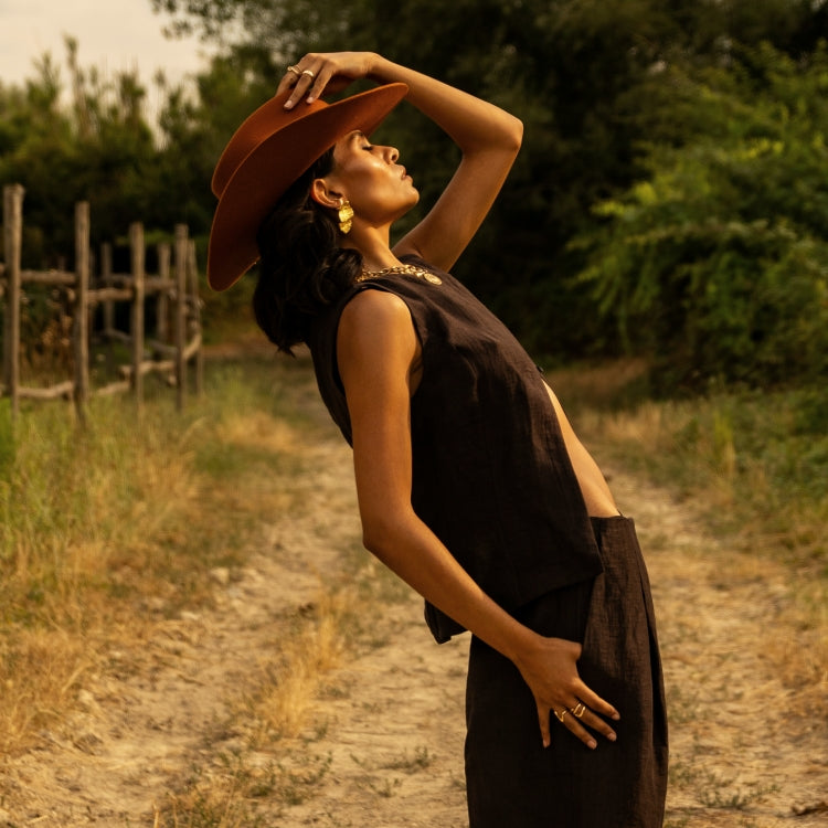 Woman wearing The Rodeo Terra Cotta — an orange wool fedora with a western-inspired crown, styled with gold statement jewelry and a sleeveless brown co-ord, photographed in golden light for Old Habits Die Hard, an Amsterdam-Based hat wear brand.