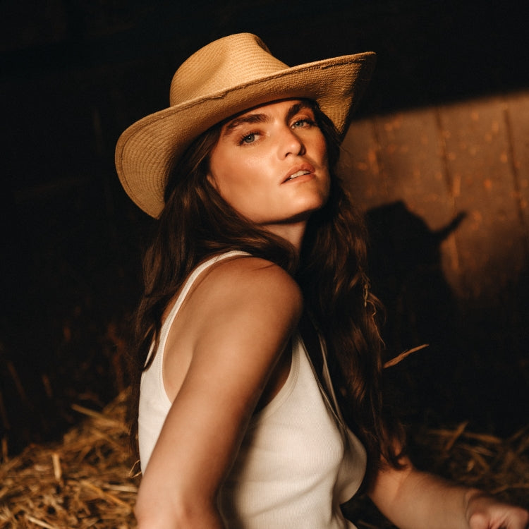 Woman wearing The Straw Habano — a neutral straw cowboy hat with a classic western shape, styled with a white tank top in golden barn light, by Amsterdam-based hatwear brand Old Habits Die Hard.