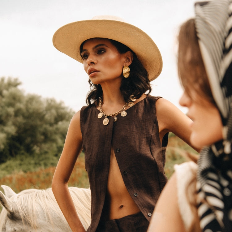 Woman wearing The Straw Sandstorm. A white straw cowboy hat with western edge, styled for summer with bold gold jewelry and a tailored brown vest, photographed for Old Habits Die Hard.