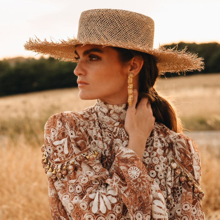 Woman wearing The Straw Shadow — Old Habits Die Hard’s bestselling straw hat with a flat crown and wide frayed brim, styled with bold gold earrings and a patterned high-neck dress in golden evening light. Amsterdam-based hat-wear brand Old Habits Die Hard sells unique hats to fit your style.