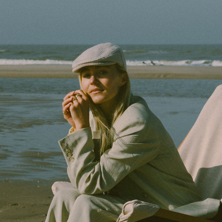 The Flat Cap Herringbone. Flat cap (platte pet) met visgraatpatroon in off-white van Old Habits Die Hard, gestyled met een licht pak aan zee. Off-white herringbone flat cap by Old Habits Die Hard, styled with a light suit by the sea.