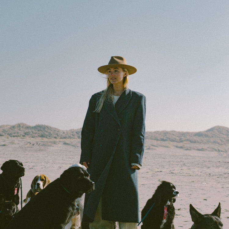 The Iconic Bombay Brown Fedora Hat. Bruine fedora hoed. Vrouw met klassieke bruine fedora hoed van Old Habits Die Hard, tussen een roedel honden op het strand.Woman wearing a classic brown fedora by Old Habits Die Hard, surrounded by a group of dogs on the beach.