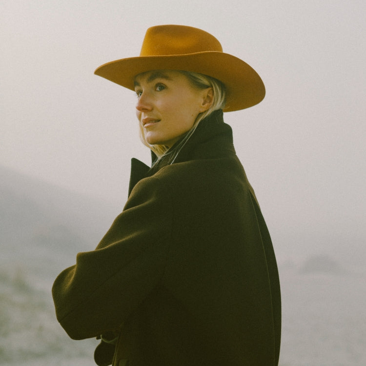 The Rodeo Terra Cotta cowboy hat. Western-geïnspireerde fedora hoed in terracotta kleur van Old Habits Die Hard, gedragen met een donkere jas in mistig landschap. Western-inspired fedora hat in terracotta by Old Habits Die Hard, styled with a dark coat in a misty landscape. Cowboy hoed voor het festival seizoen