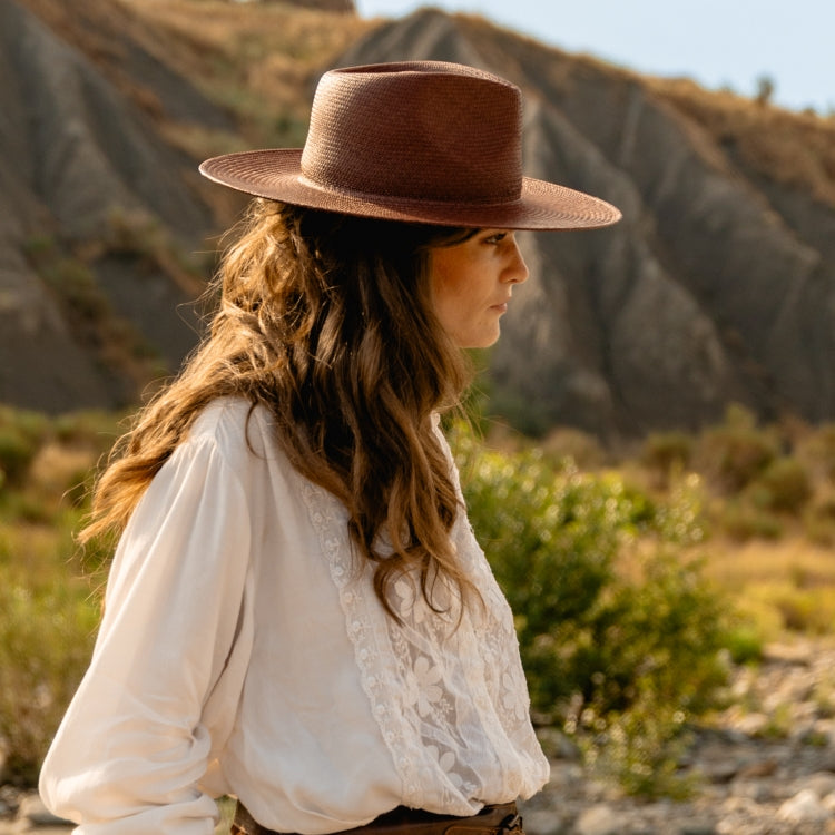 Woman wearing The Straw Iconic Glowy Bronze — a brown straw fedora with a flat crown and structured brim, styled with a white embroidered blouse against a rugged mountain backdrop, by Old Habits Die Hard, an Amsterdam-Based hatwear brand.
